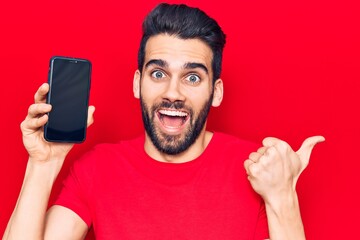 Young handsome man with beard holding smartphone showing screen pointing thumb up to the side smiling happy with open mouth