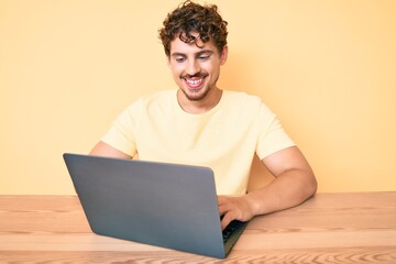 Young caucasian man with curly hair working at the office with laptop looking positive and happy standing and smiling with a confident smile showing teeth
