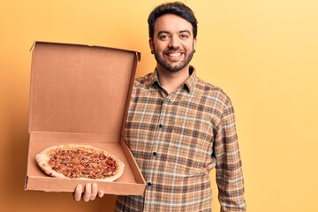 Young hispanic man holding delivery pizza box looking positive and happy standing and smiling with a confident smile showing teeth