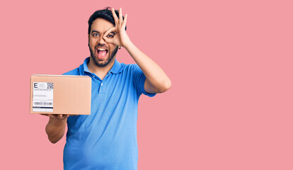 Young hispanic man holding delivery package smiling happy doing ok sign with hand on eye looking...