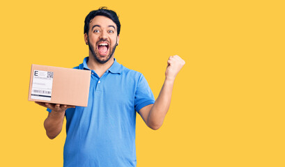 Young hispanic man holding delivery package screaming proud, celebrating victory and success very...