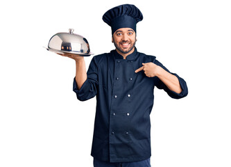 Young hispanic man wearing cooker uniform holding tray pointing finger to one self smiling happy and proud