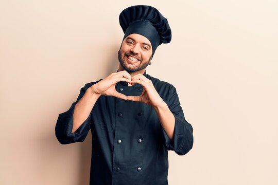 Young hispanic man wearing cooker uniform smiling in love doing heart symbol shape with hands. romantic concept.