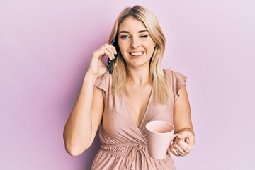 Young caucasian woman talking on the smartphone and drinking a cup of coffee winking looking at the...