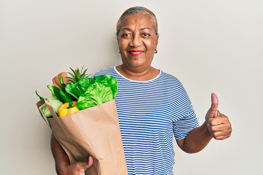 Senior african american woman holding paper bag with bread and groceries smiling happy and positive, thumb up doing excellent and approval sign