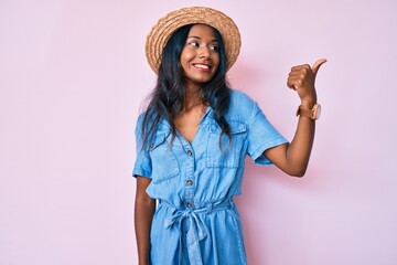 Young indian girl wearing summer hat pointing thumb up to the side smiling happy with open mouth