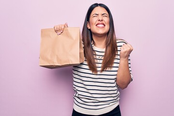 Young beautiful woman holding deliver paper bag with takeaway food over pink background screaming...