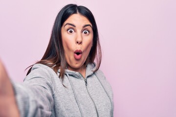 Young beautiful brunette sporty woman make selfie by the camera over pink background scared and amazed with open mouth for surprise, disbelief face