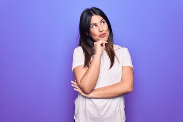 Young beautiful brunette woman wearing casual t-shirt standing over purple background thinking concentrated about doubt with finger on chin and looking up wondering