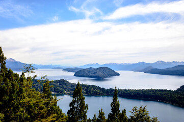 Obraz premium Beautiful lake landscape in the middle of mountains surrounded by pine trees and rocks everywhere. Very sunny day in the mountain of Bariloche. Green foliage and huge rocks.