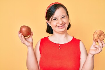 Brunette woman with down syndrome holding red apple and donut sitting winking looking at the camera with sexy expression, cheerful and happy face.