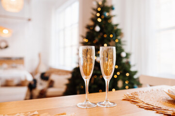 two gilded glasses for drinks on table, light cozy Scandi room with large Christmas tree and lights on background. selective focus