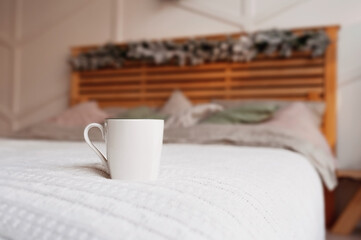 clear white ceramic mug on cozy wooden bed in christmas decor with bedspread of pastel colors, pillows of powdery shades and blanket of delicate colors. Stylish bedroom in Scandi style,