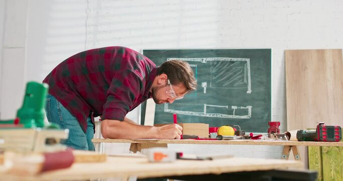 A handsome carpenter in a flannel shirt counts the dimensions on a smartphone in a carpentry workshop. A bearded handyman wearing protective glasses checks the design on a sheet of paper. In the