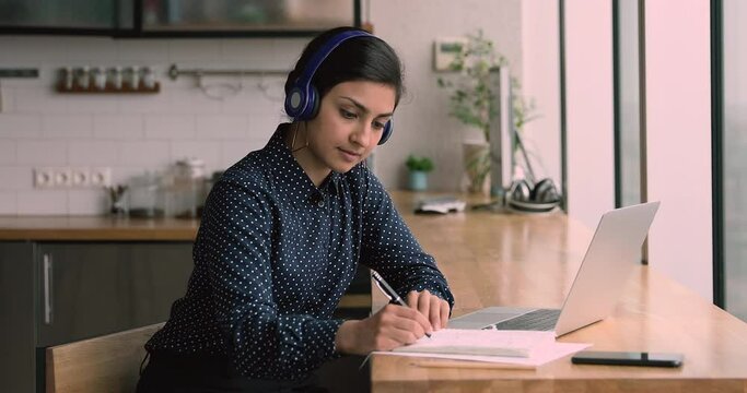 Indian millennial woman sit table in domestic kitchen wear headphones listen audio course english learn language using laptop and internet, writing notes memorizing information. Self-education concept