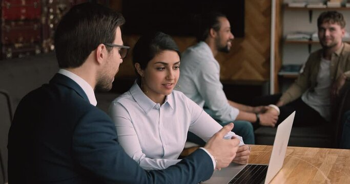In modern co working office talking diverse colleagues indian businesswoman and confident businessman in formal suit, workmates sit at desk use laptop, discuss wok result, new business e app concept