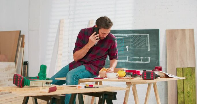 A carpenter uses a smartphone during a break at work. Young handsome craftsman is resting in a carpentry workshop. The young man is talking on the phone. There are a locksmith table and tools in the