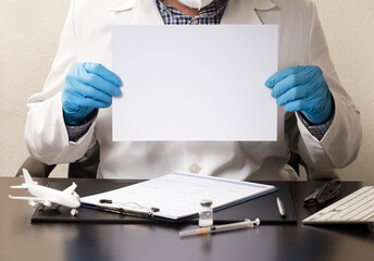 Doctor with blue gloves, white poster in hands (to fill out), desk with syringe, vaccine, airplane, glasses, white form and computer keyboard