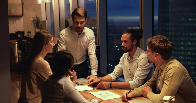 Five multi ethnic young startupper diverse team working until late in modern office, sit at table in illuminated boardroom discuss sales stats financial results engaged in group brainstorming concept