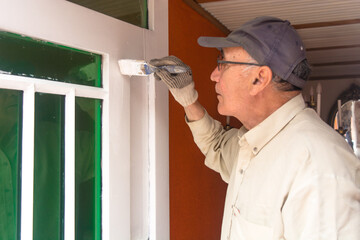 Naklejka na ściany i meble Old man with beige shirt and grey cap, holding a brush with white paint and painting a door with green windows