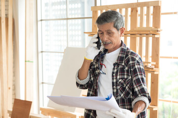 Fototapeta premium Portrait Senior asian elderly old man carpenter wearing white gloves protection using phone mobile to make a call and holding blueprint plan in workshop