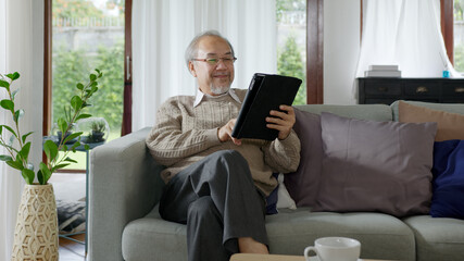 Candid of old asian retired man using tablet computer to transfer money online or financial payment and read text, smile or laugh with happy and positive. Senior asian with digital technology concept