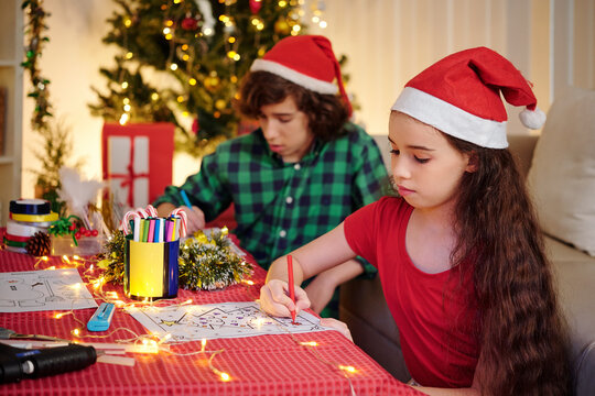 Hispanic siblings in Santa Claus hats concentrated on drawing Christmas pictures