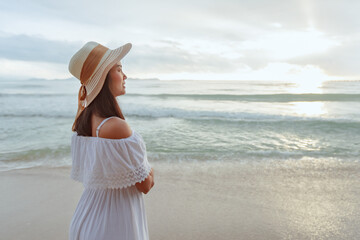 Fototapeta na wymiar Asian woman wearing stylish hat and clothes looking to the ocean and enjoying beautiful Sunset on the Beach. Summer holidays and travel concept.