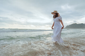 Fototapeta na wymiar Asian woman wearing stylish hat and clothes walking on the beach and enjoying beautiful nature in the sunset time. Summer holidays and travel concept.