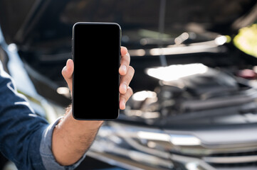 Male hand showing blank screen of smartphone. In front of broken down car. Emergency assistance service