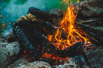 Vivid smoldered firewoods burned in fire close-up. Atmospheric background with orange flame of campfire and blue smoke. Warm full frame image of bonfire with glowing embers in air. Bright sparks bokeh