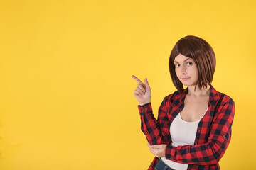 A young brunette girl with short hair in a checkered red shirt and jeans points to an empty space for text on a yellow background and smiles. Half body