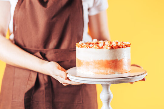 Woman Baker Holding Cake On A Stand. Uniforms Of Kitchen Workers In Front Of A Yellow Background. Sweets For Any Holiday