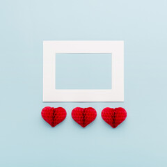 Valentine's Day holiday card. Red small paper hearts and white empty frame on pastel blue background. Valentines day concept.