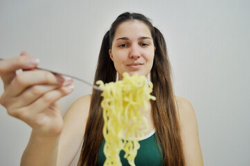 Young girl with dark hair eating instant noodles with a fork on light background