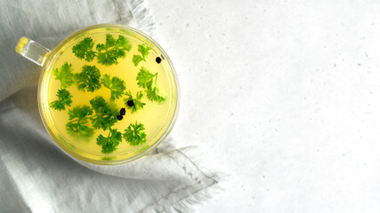Cup of homemade chicken bone broth on white background with textile. Parsley leaves and peppercorns. Health benefits. Collagen is good for joints. Flat lay. Copy space. Horizontal orientation.