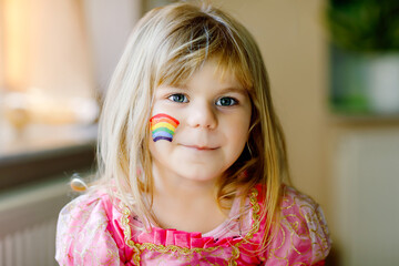 Portrait of cute little toddler girl with painted rainbow with colorful colors on face. Lonely child during pandemic coronavirus quarantine. Children make and paint rainbows around the world