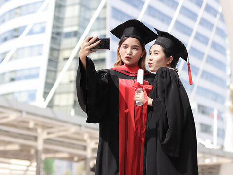 happy graduated women in graduation gowns making  kissing lips, taking selfie photo with mobile phone. City building background. Education, Friendship and technology concept..
