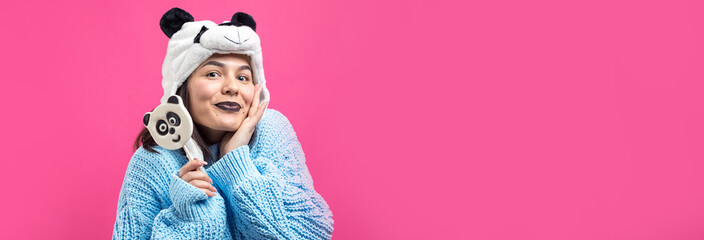 Funny young girl standing with yummy panda-lollipop in her hand and a hat on his head on a pink background.