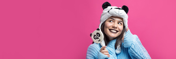 Funny young girl standing with yummy panda-lollipop in her hand and a hat on his head on a pink background.