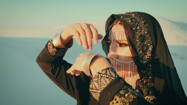 portrait of beautiful young woman in black oriental abaya dress posing in desert on white sand. head is covered with scarf, silk hijab, face is hidden by golden veil mask. Girl dancing with her hands 