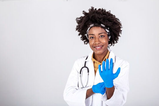 Portrait of an attractive young female doctor in white coat putting on medical gloves. Portrait of glad smiling doctor in white uniform standing with crossed hands on gray background