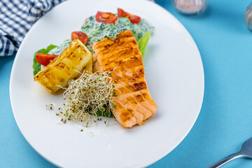 Grilled salmon steak. A piece of red fish on a white plate and a blue background on the table. Fish food with sauce, tomatoes, lemon. Restaurant menu. Grill fried.
