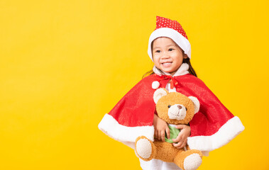 Kid dressed in Santa Claus hat, Portrait of Asian little cute girl smiling hugging teddy bear the concept of Christmas Xmas day, studio shot isolated on yellow background with copy space for text
