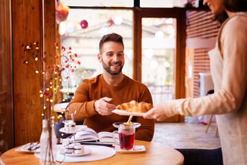 Happy casual man having breakfast in the cafe. Smiling guy sitting at desk while waitress bringing croissant to him in cafeteria. 