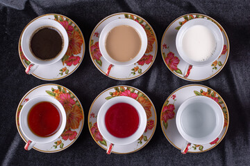 Six cups and saucers filled with water, juice, tea, coffee, milk and coffee with milk on a dark fabric background © Aleksey