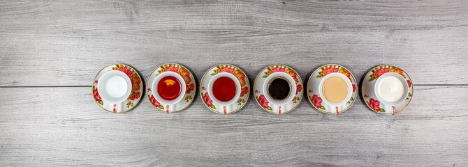 Six cups and saucers on a wooden table filled with water, juice, tea, coffee, milk and coffee with...