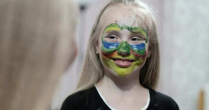 Schoolgirl with face painting looks in the mirror. A cute girl with a multi-colored face is smiling. Body art.