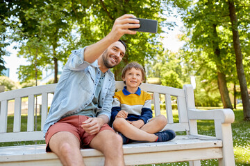 Fototapeta na wymiar family, fatherhood and people concept - happy father and his little son taking selfie with smartphone sitting on bench at summer park