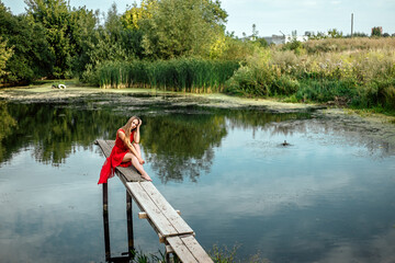Fototapeta na wymiar Full length portrait of young pretty woman sitting on the wooden pier near the river or lake touching clothes, dressed in red dress, looking at the camera, copy space and nature blur background.